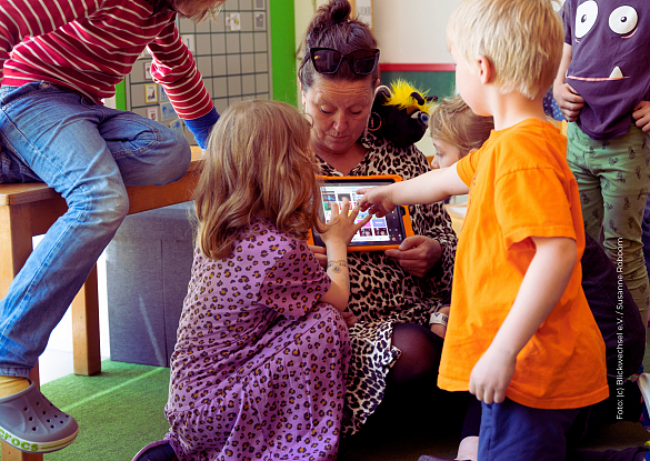 Erzieherin, die ein Tablet in der Hand hält und alle Kinder um sie herumstehen und auf das Tablet schauen